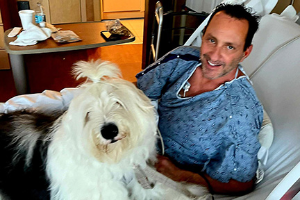 Scott smiles while recovering in his hospital room with his white dog sitting on his lap, sharing a comforting moment together.